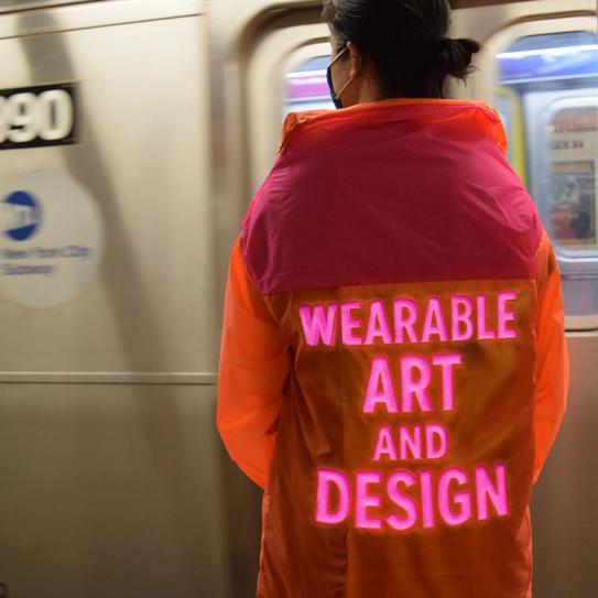 person from the back wearing a vibrant orange and pink jacket. features glowing neon pink text that reads 'WEARABLE ART AND DESIGN'