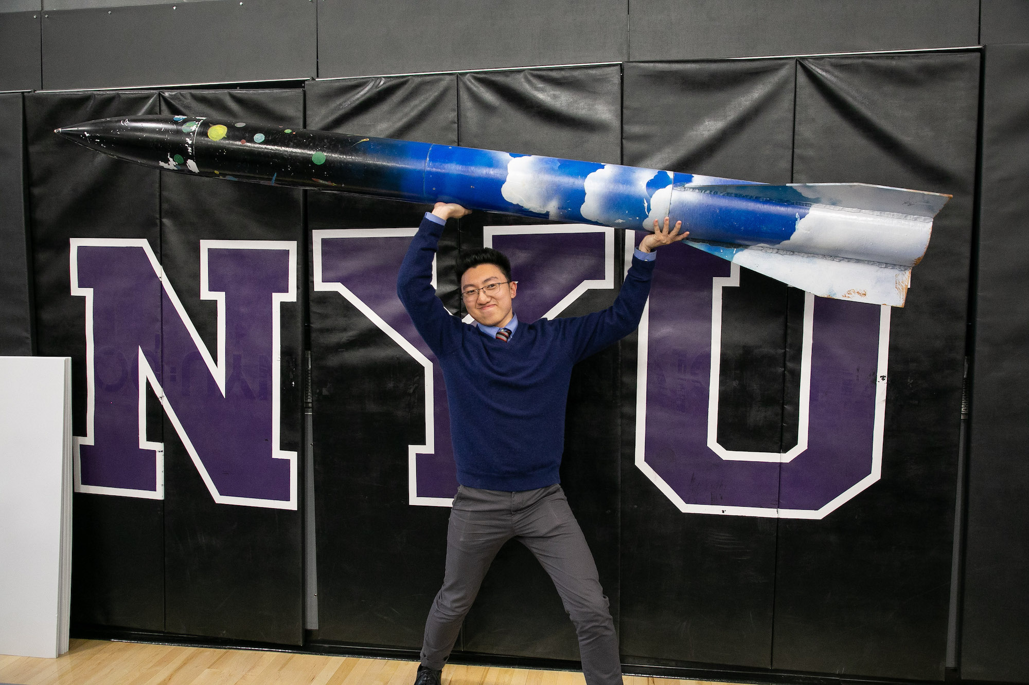 male student holding a rocket overhead in front of NYU banner